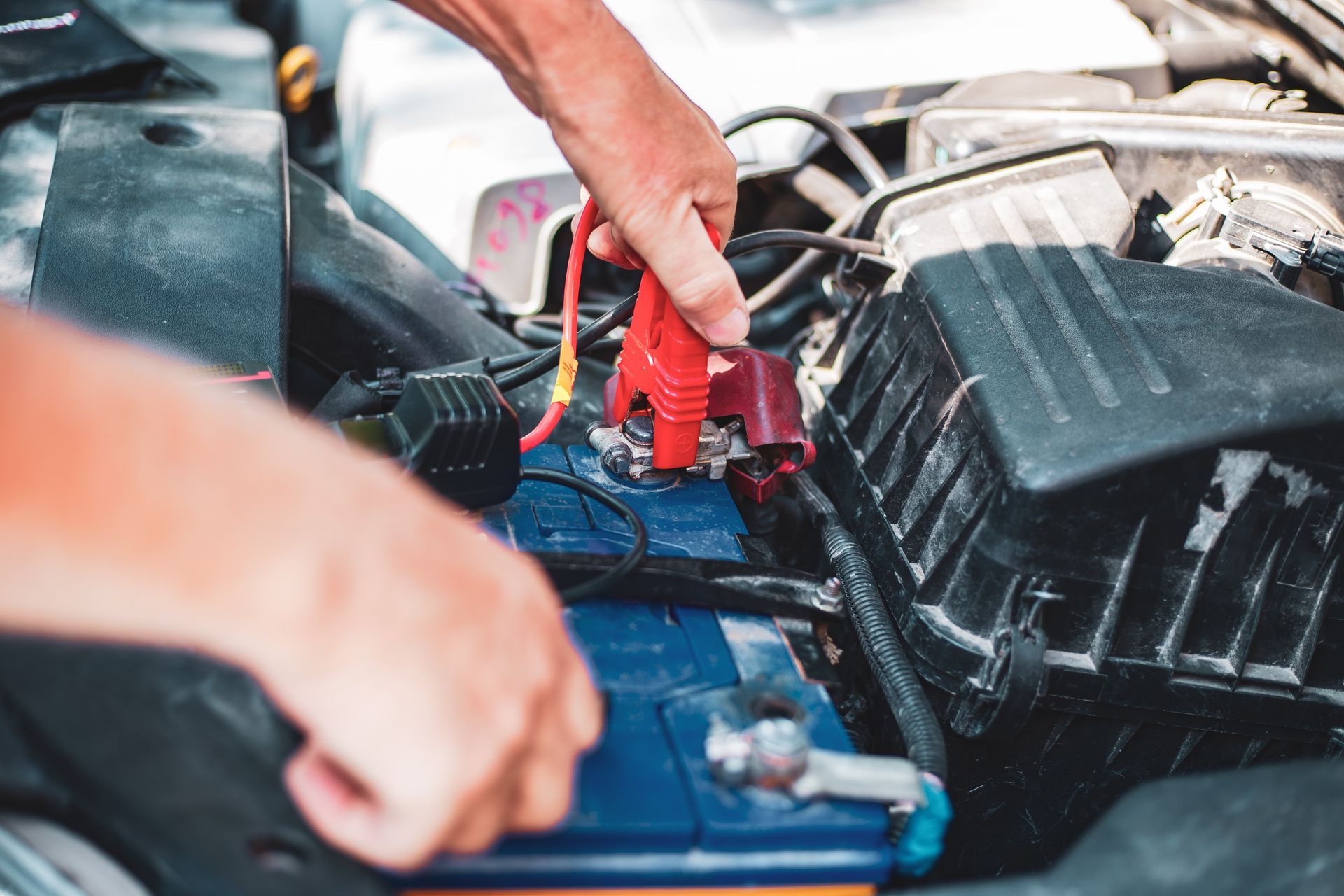 Hands connecting a red jumper cable to a car battery's positive terminal in an engine bay.