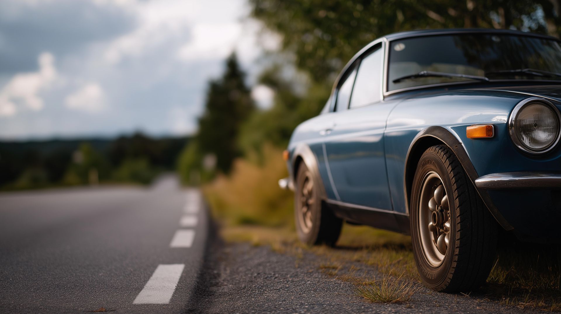 Blue classic car parked on the side of a road, blurred trees and sky in the background.