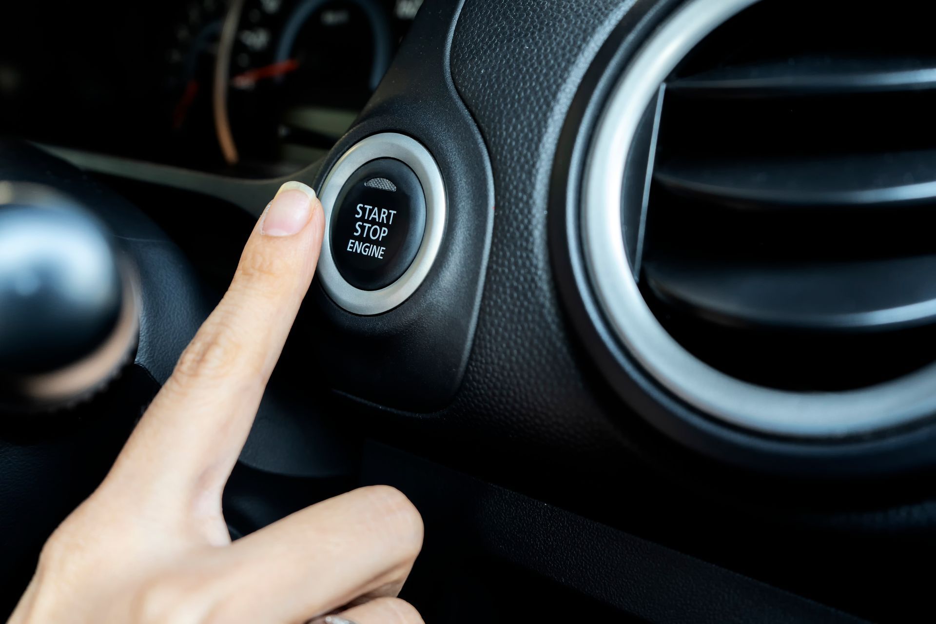 Finger pressing a black car's start/stop engine button, next to an air vent.