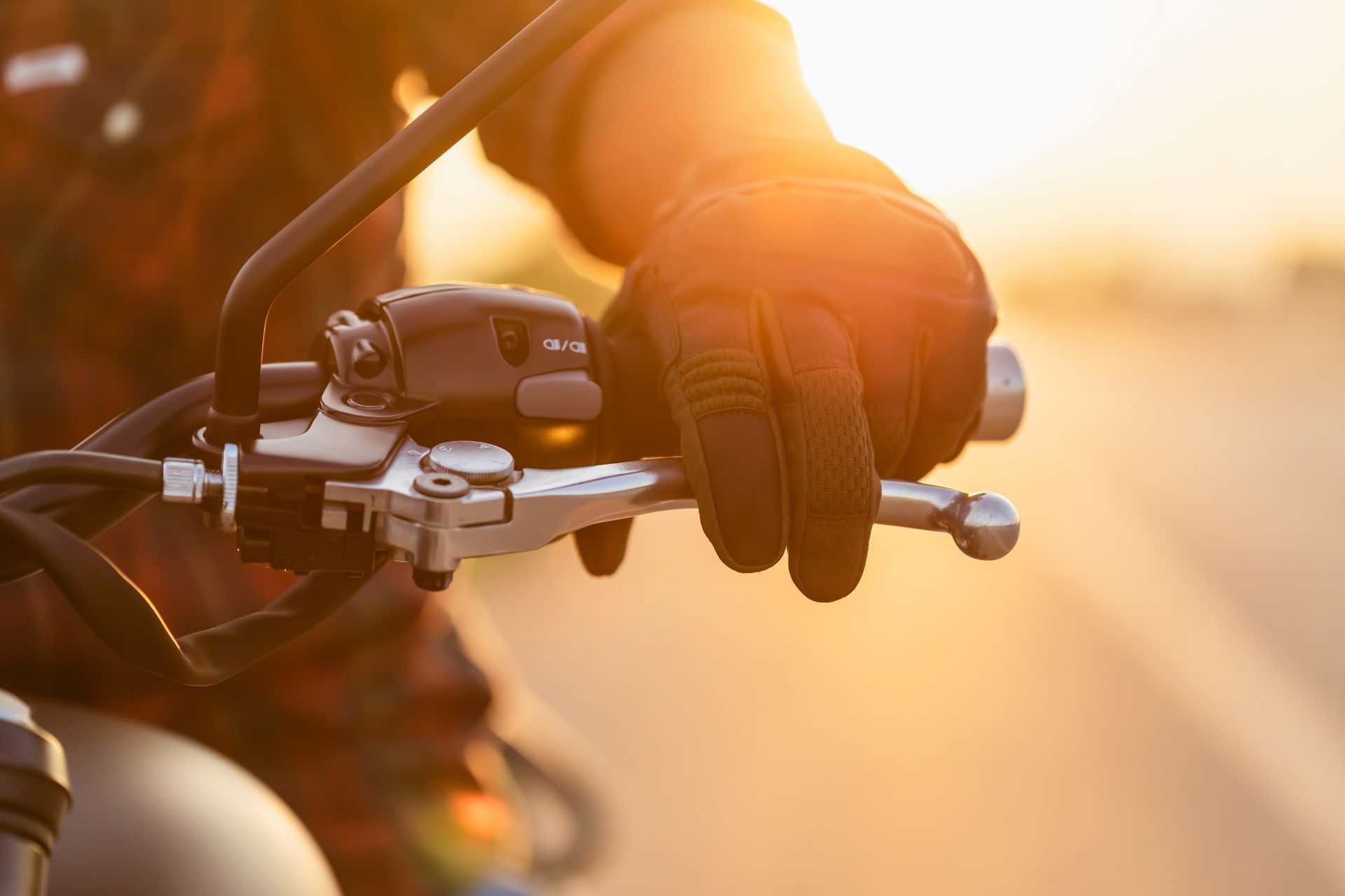 Hand wearing a black glove grips a motorcycle handlebar; sunlight bathes the scene.