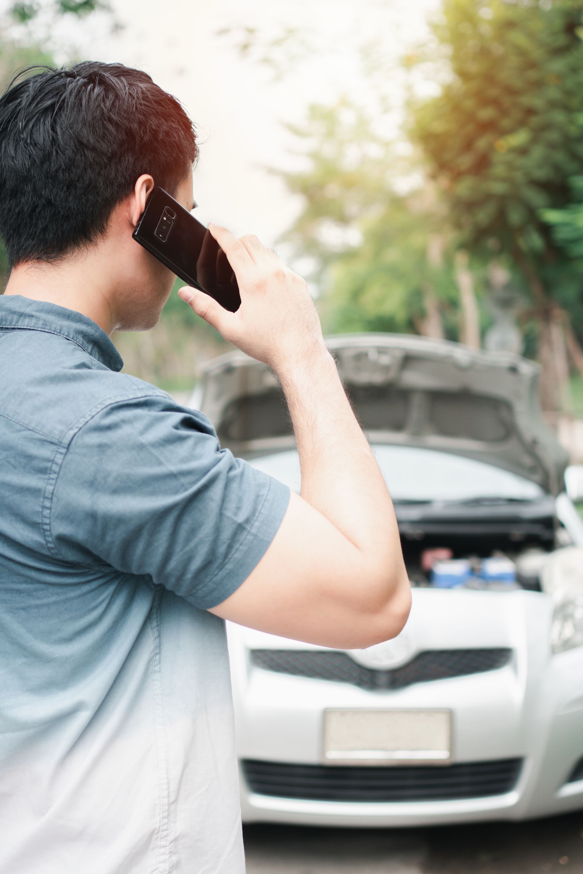 Man on phone with a broken-down car, hood up, outdoors.