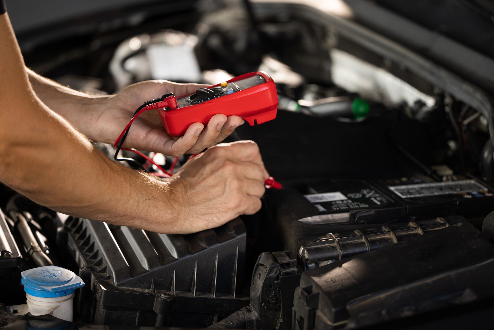 Hands holding red multimeter, testing car battery in an open engine bay.
