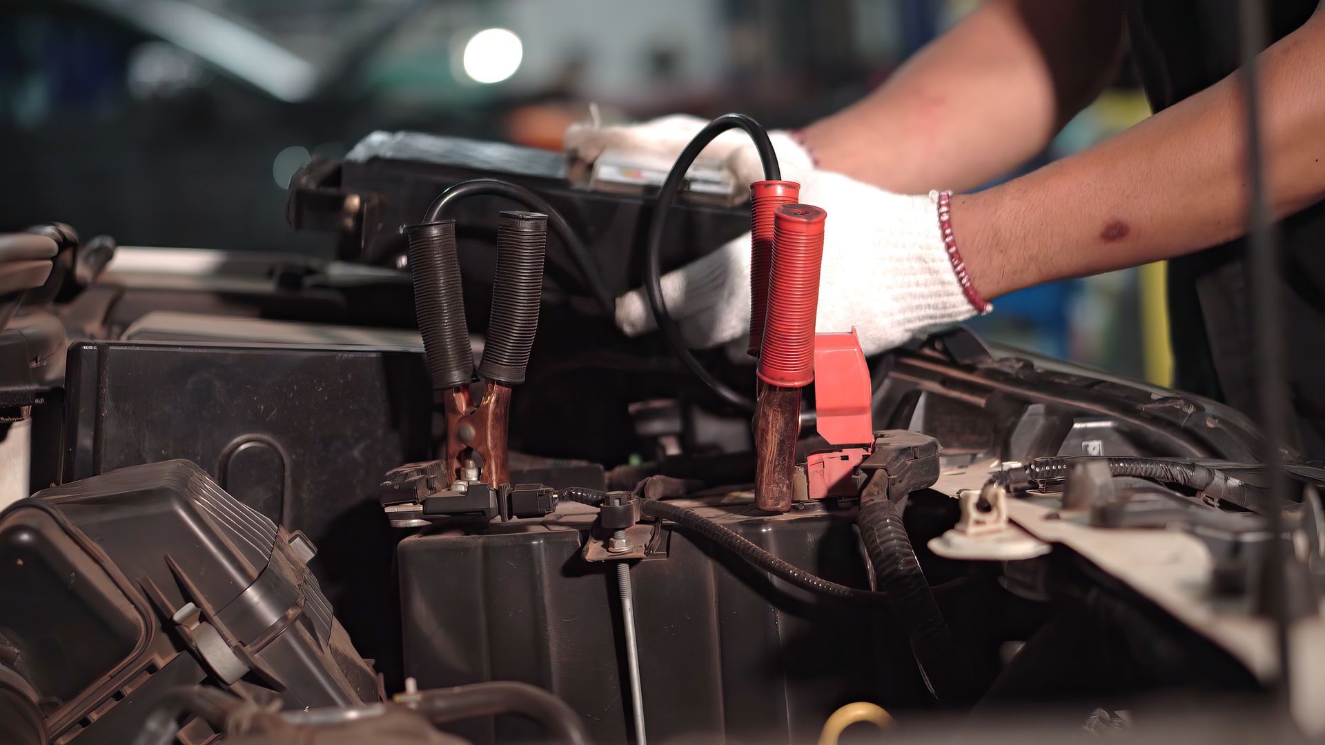 Hands in gloves connecting jumper cables to a car battery in a dimly lit setting.