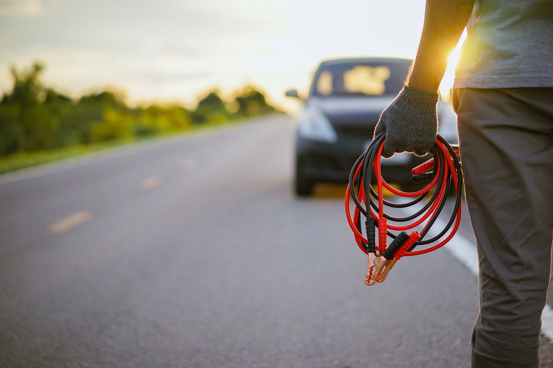 Person holding jumper cables near a car on a road, with sunlight.