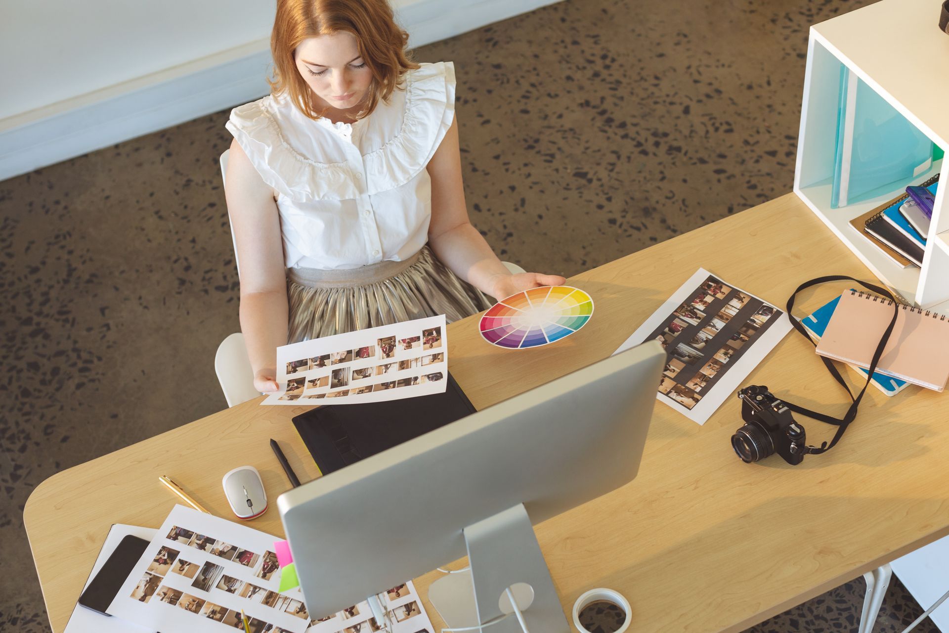 Woman at desk, holding photos and a color palette, working on a computer.