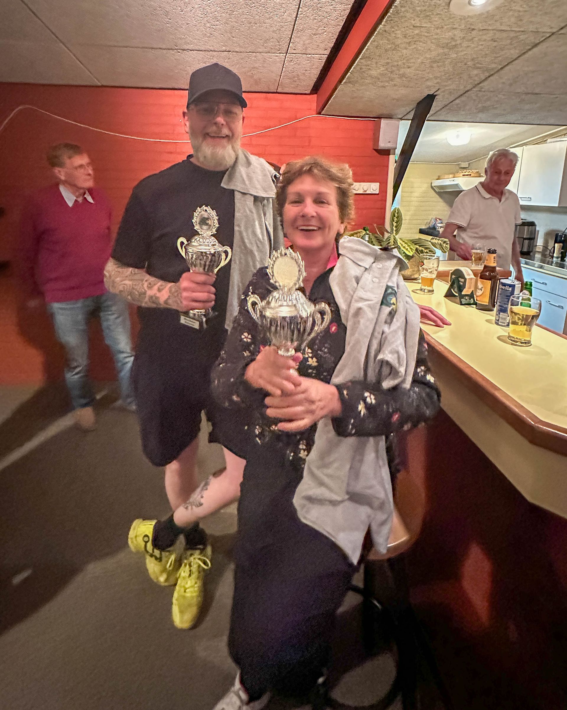 Twee mensen met trofeeën in hun handen aan de bar, lachend. Een man in een zwart shirt, een vrouw in een blouse met bloemenprint. Anderen staan ​​achter de bar.