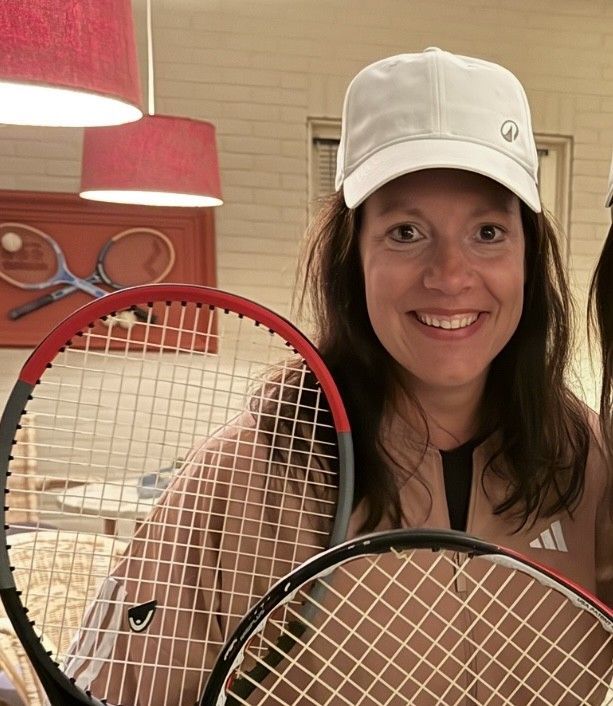 Een lachende vrouw met tennisrackets in haar handen en een witte pet op. Roze en zwarte outfit. Interieur.