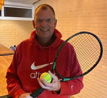 Een man in een rode hoodie met een tennisracket en -bal staat lachend voor een bakstenen muur.