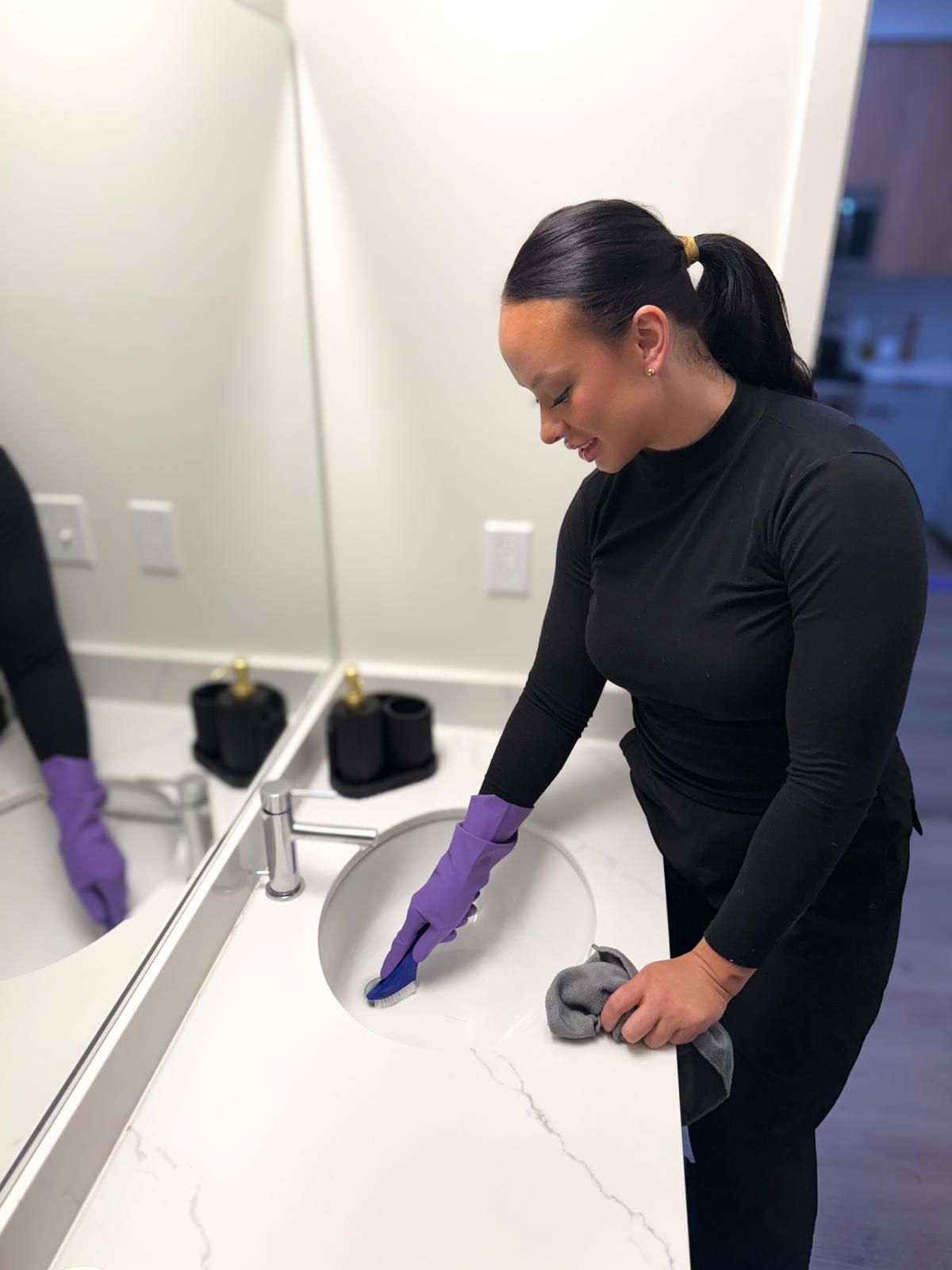 Person wearing purple gloves cleans a white bathroom sink with a brush and cloth.