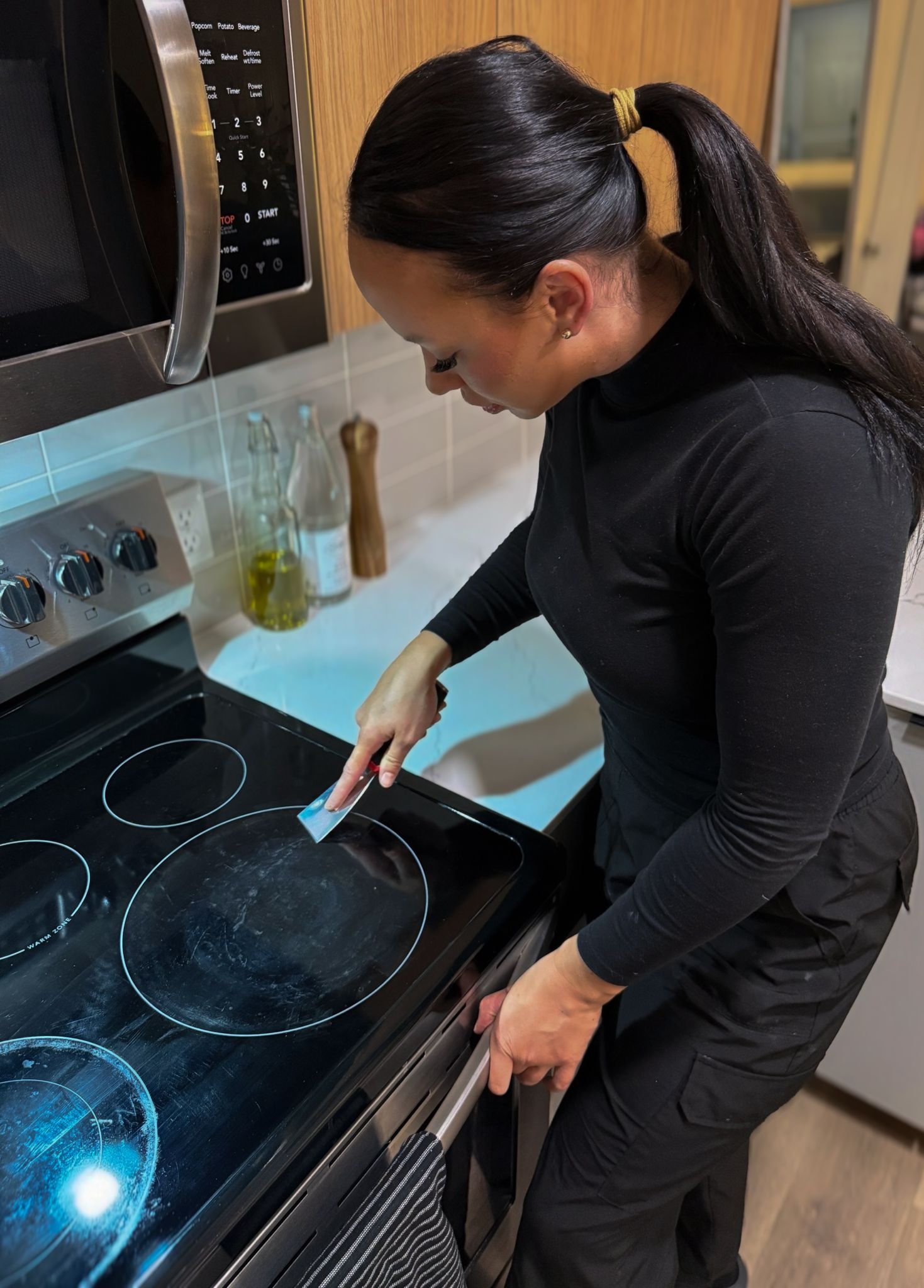 Woman cleaning stovetop in a kitchen; she's wearing black and using a scraper.