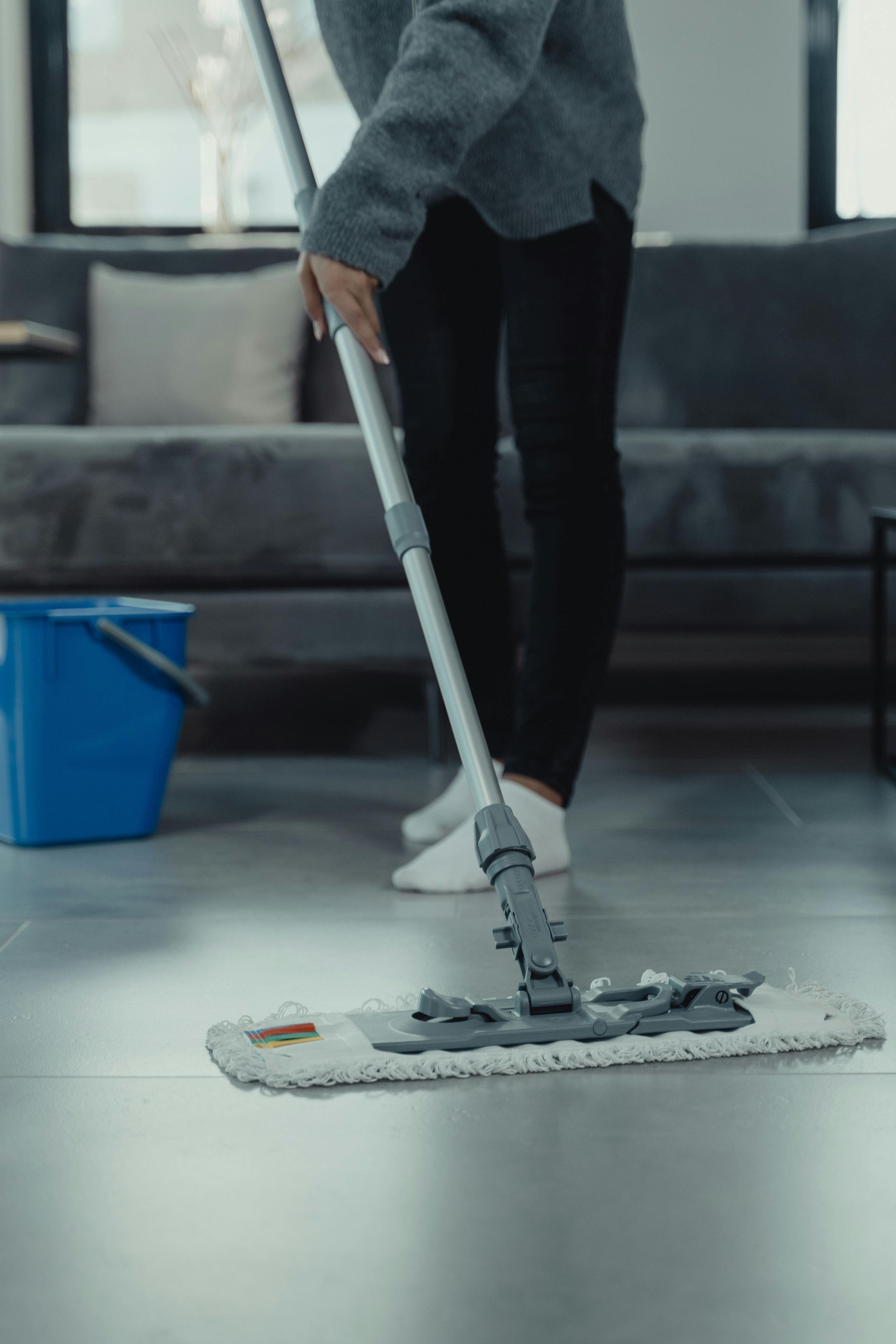 Person mopping a gray tile floor in a living room, blue bucket to the side, gray sofa in background.