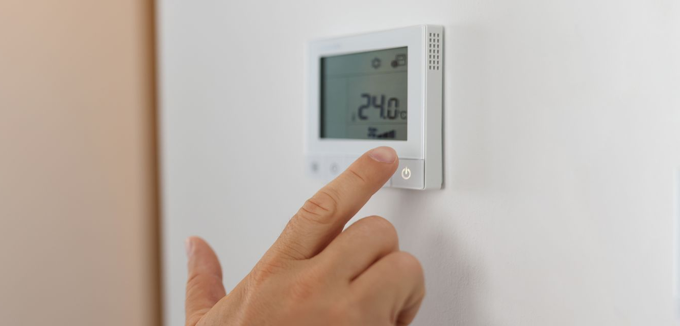 La main d'une personne appuie sur un bouton d'un thermostat blanc affichant 24,0.