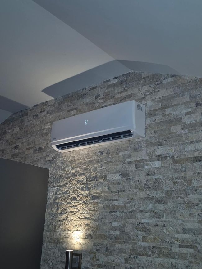 Climatiseur monté sur un mur en pierre texturée. Un spot en dessous éclaire le mur.