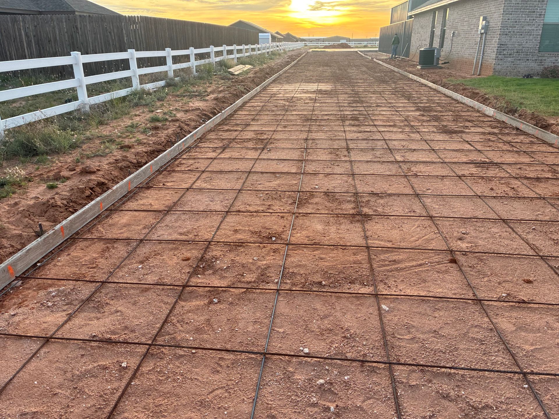 Brick-paved driveway under construction at sunset, bordered by a white fence and stone buildings.