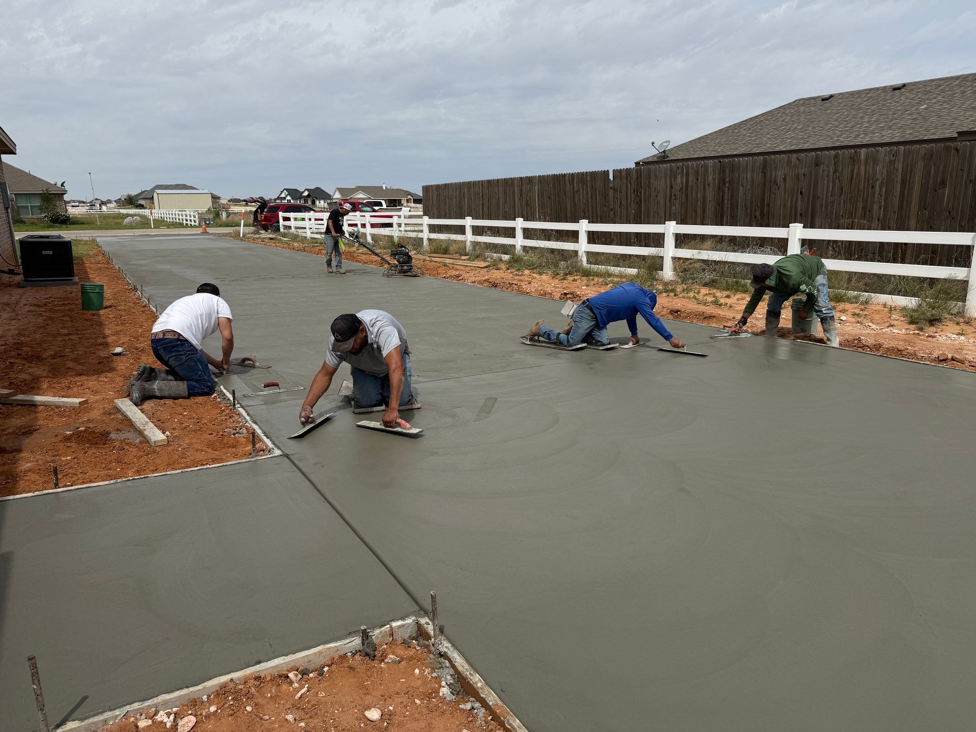 Workers smoothing fresh concrete on a driveway beside a white fence and homes