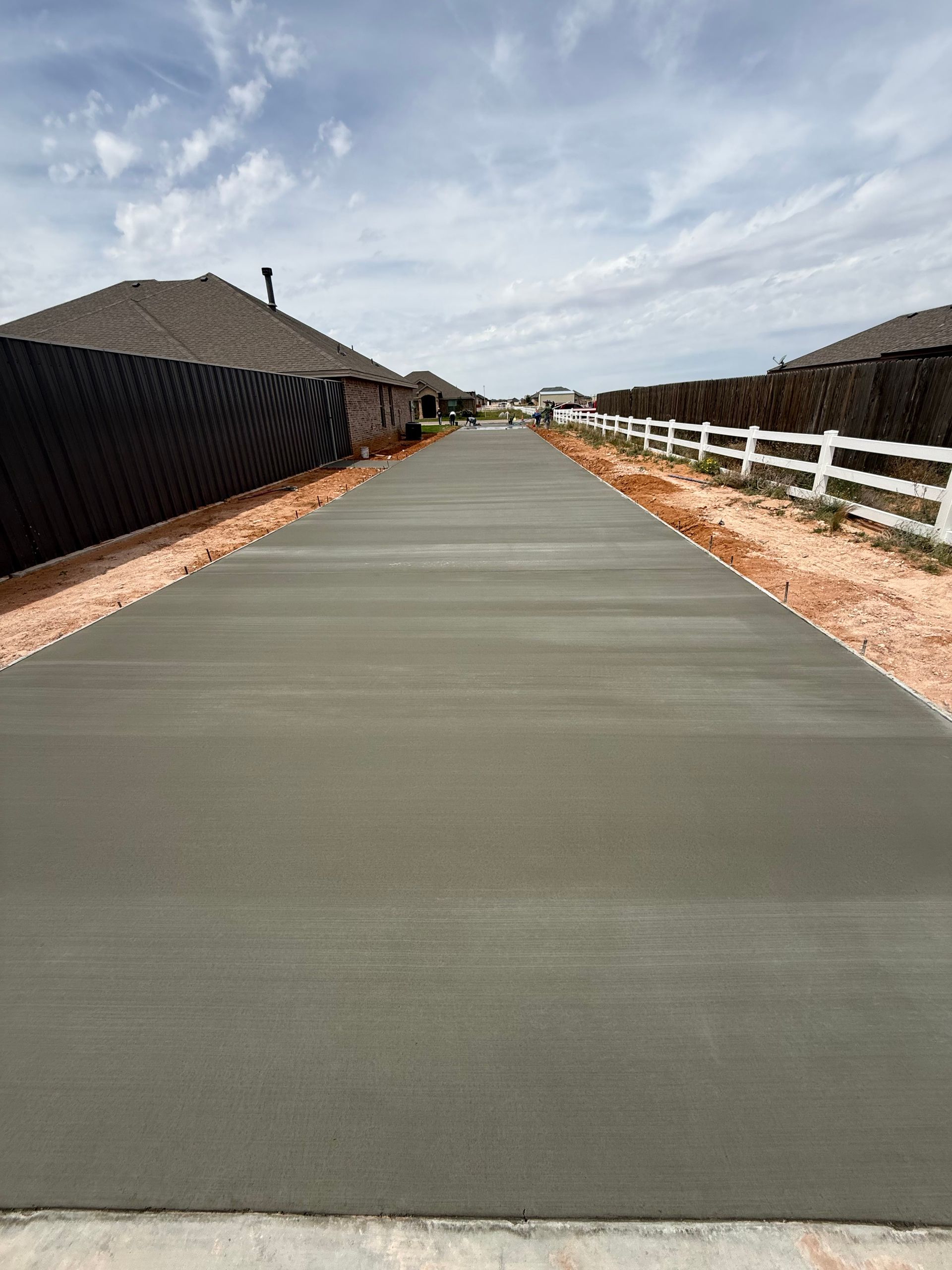 Freshly poured concrete driveway between fenced suburban houses under a cloudy sky