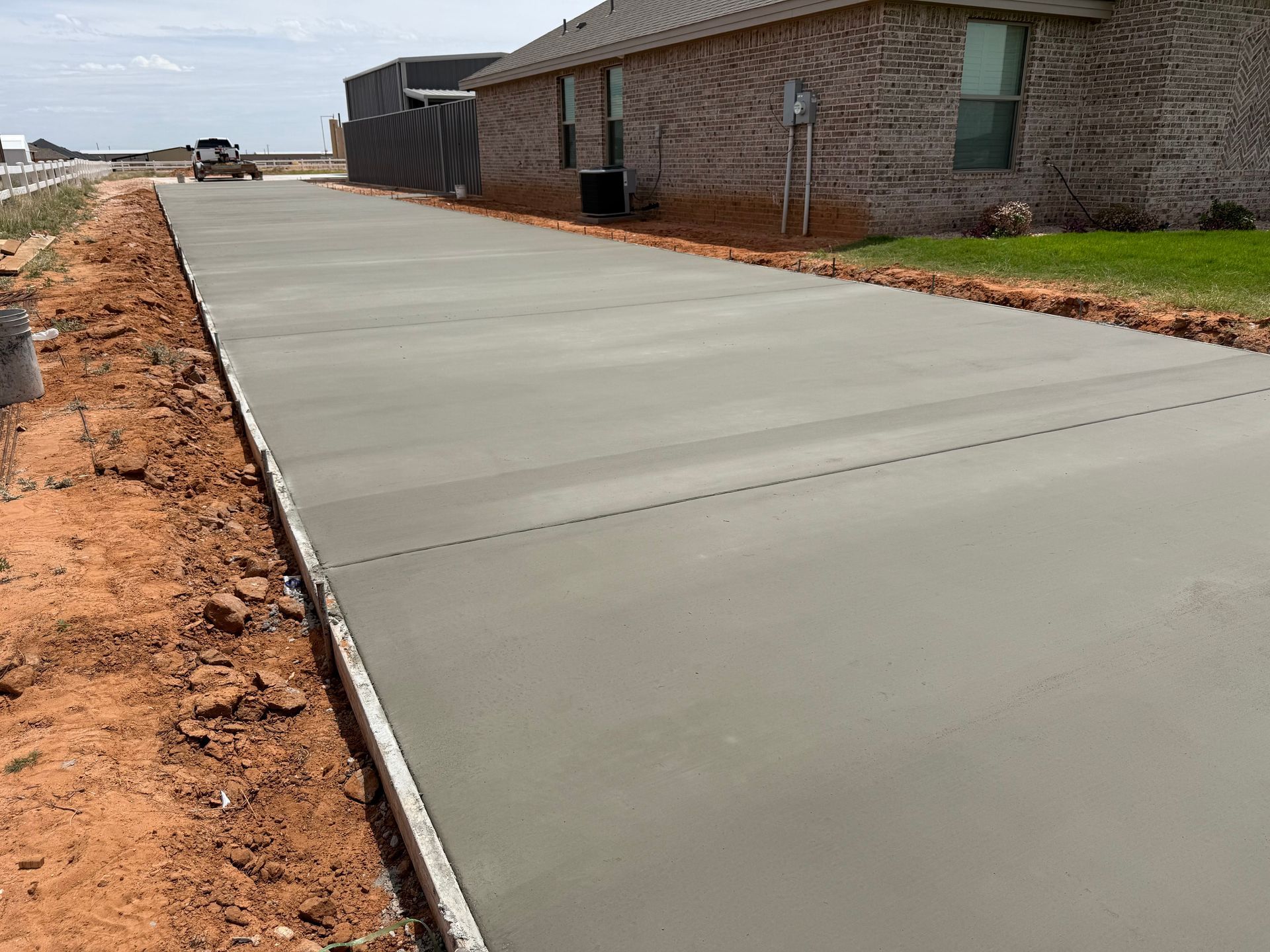 New concrete driveway beside a brick house, bordered by dirt and construction materials