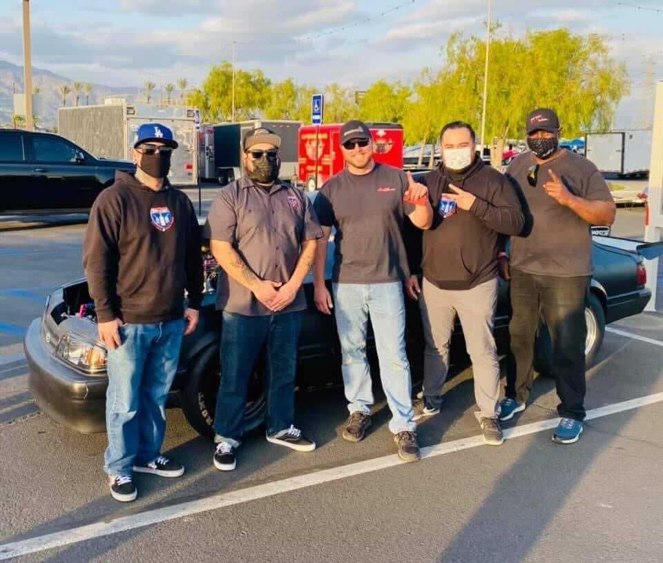 Five people pose in front of a car with its hood open, outdoors. All wear masks.