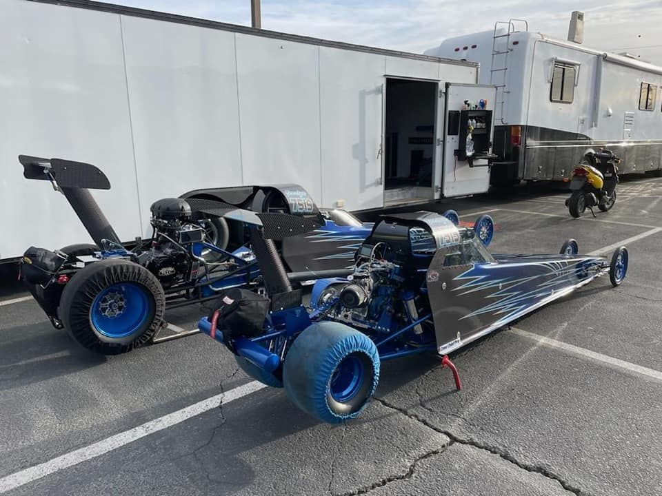 Two blue dragsters parked on asphalt, near a trailer.
