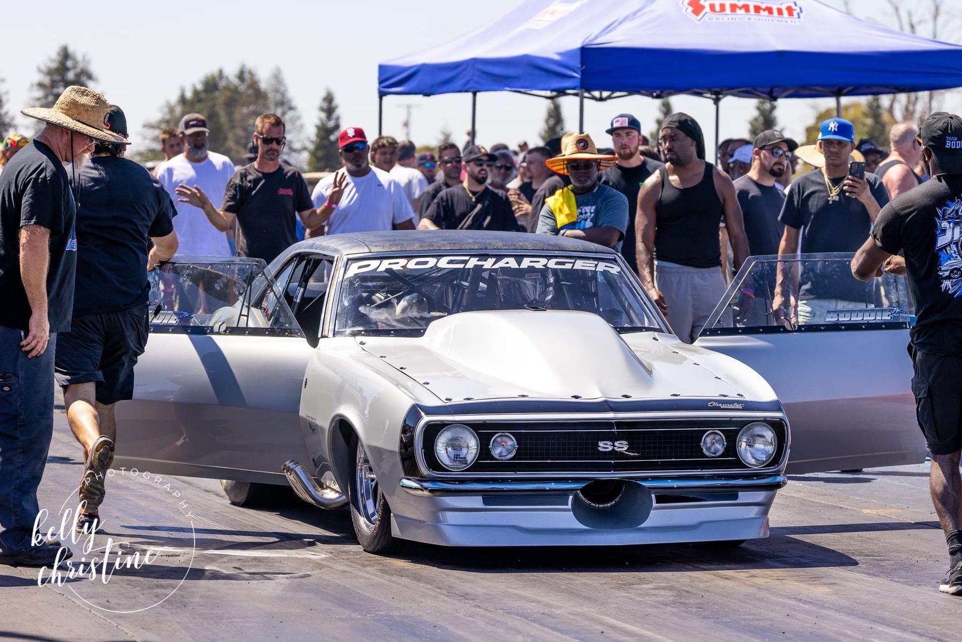 Silver race car at a drag strip, surrounded by people; blue canopy in the background.