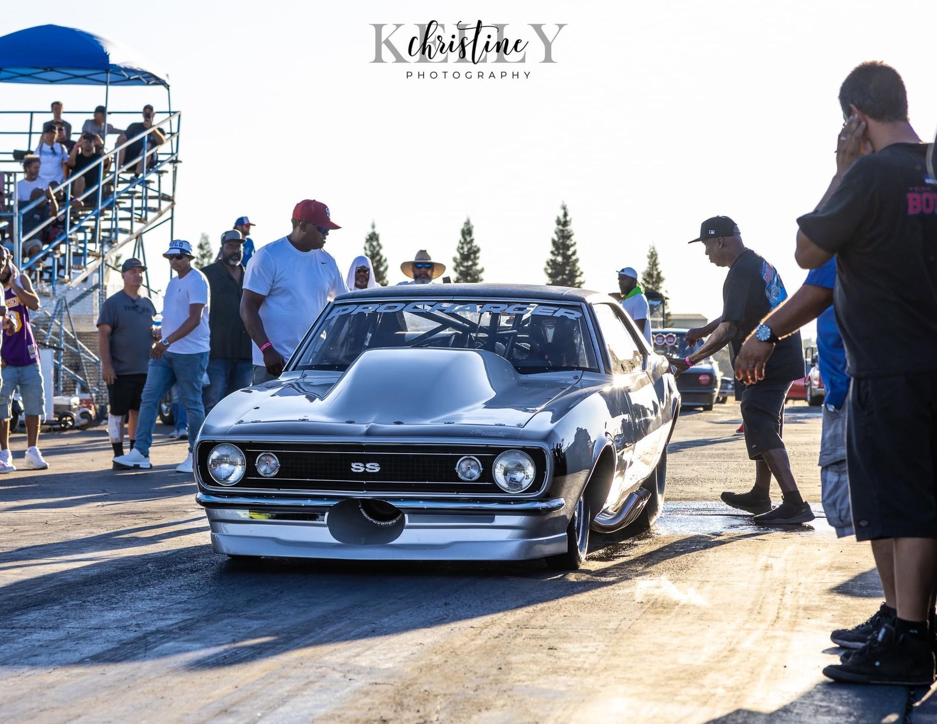 Silver classic car at a racetrack, surrounded by people.