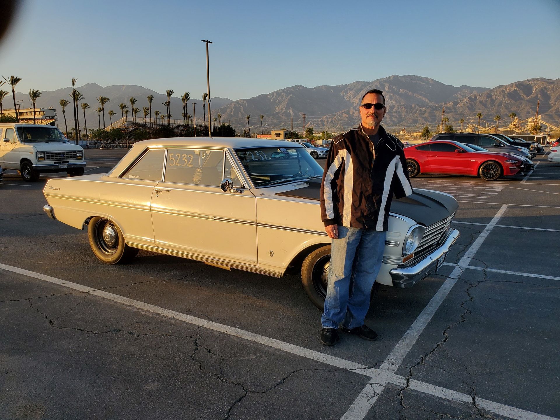 Man standing next to a classic white car in a parking lot. Mountains in the background, sunny day.