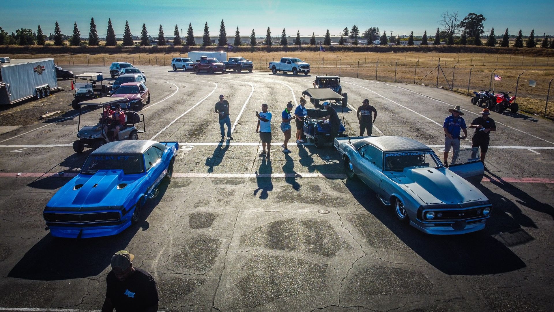 Two muscle cars staged at drag strip, surrounded by crew and spectators. Blue and silver cars, sunny day.