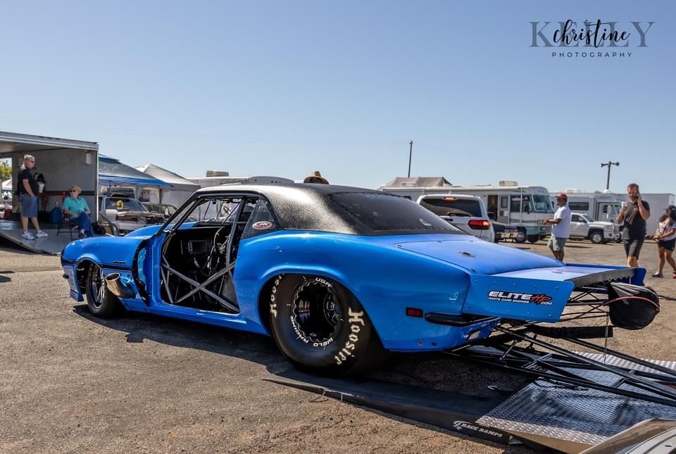 Blue race car on a trailer at a track, open doors, roll cage, slick tires.