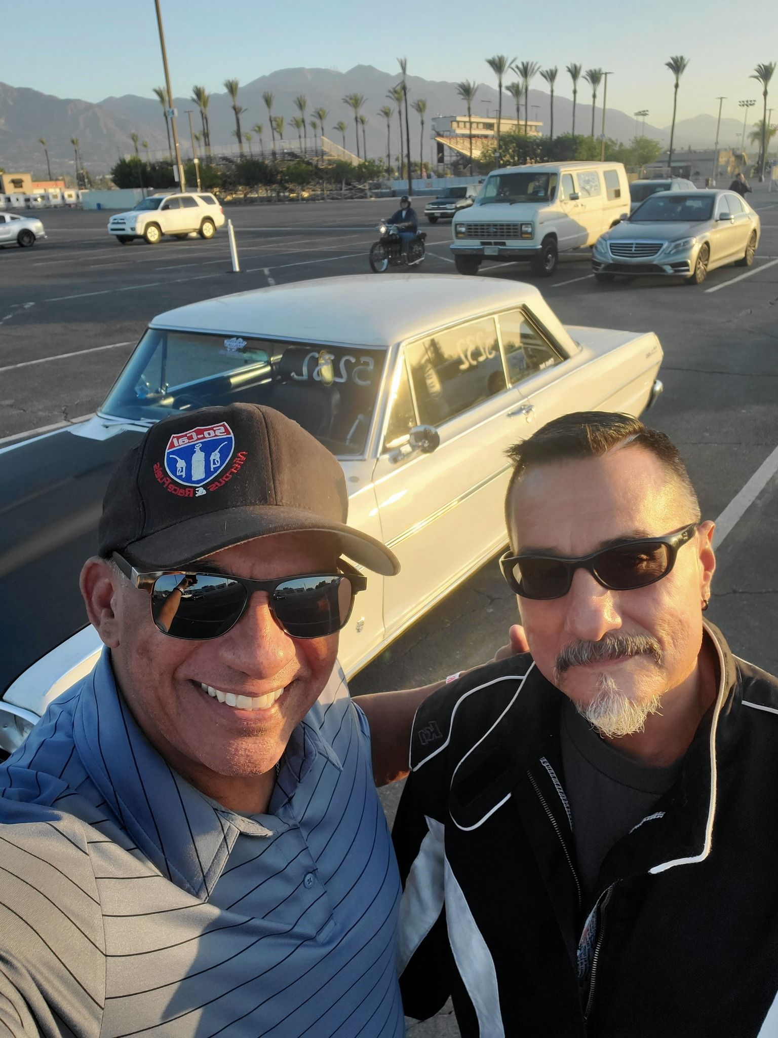 Two men smiling in front of a classic white car; palm trees and mountains in background.