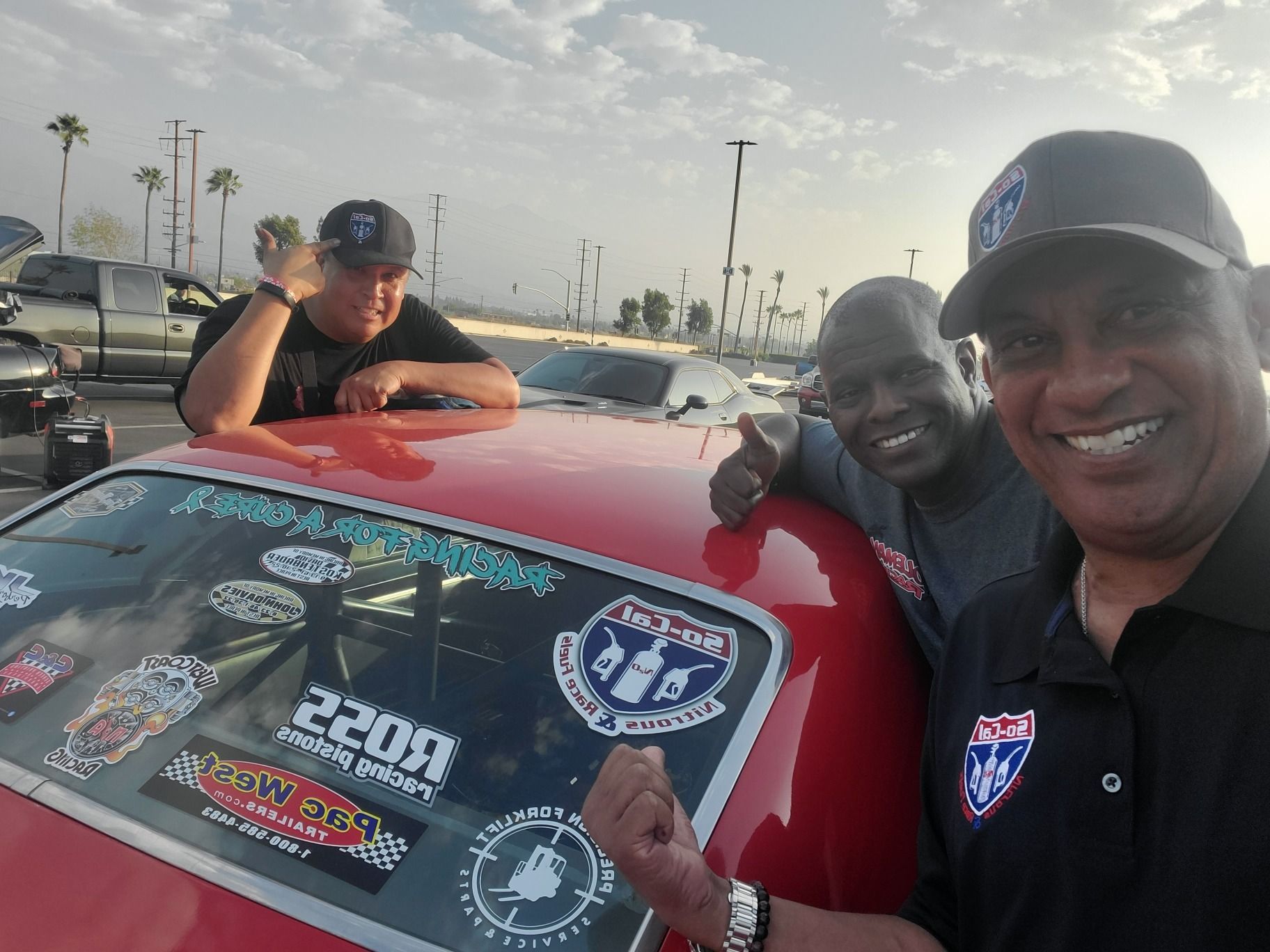 Three men pose with a red race car, thumbs up, smiles, wearing hats and shirts with logos at a track.