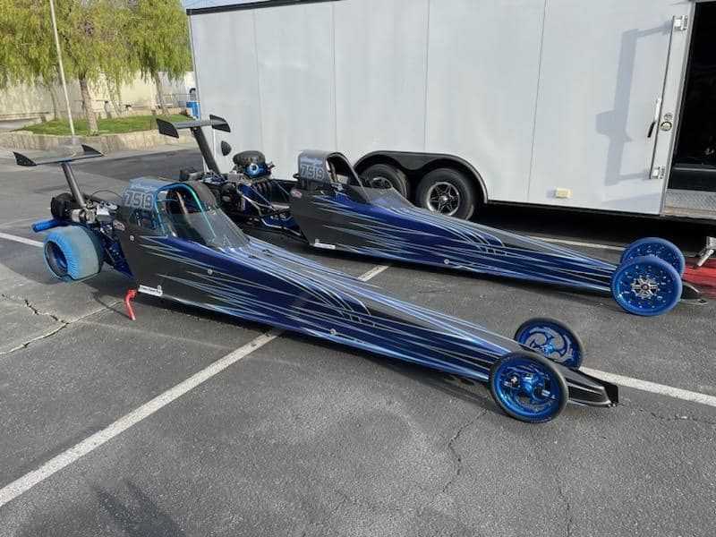 Two blue drag racing cars parked on asphalt next to a trailer.