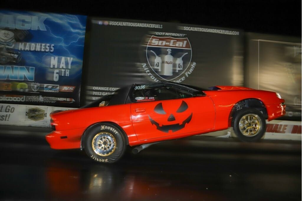Orange race car with a jack-o'-lantern design, racing at night with event signage in the background.