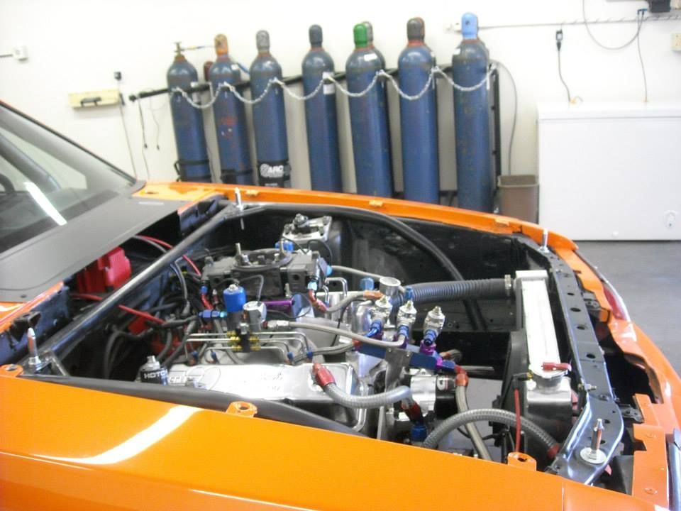 Orange race car engine bay with engine, fuel lines, radiator, and a row of gas cylinders in the background.