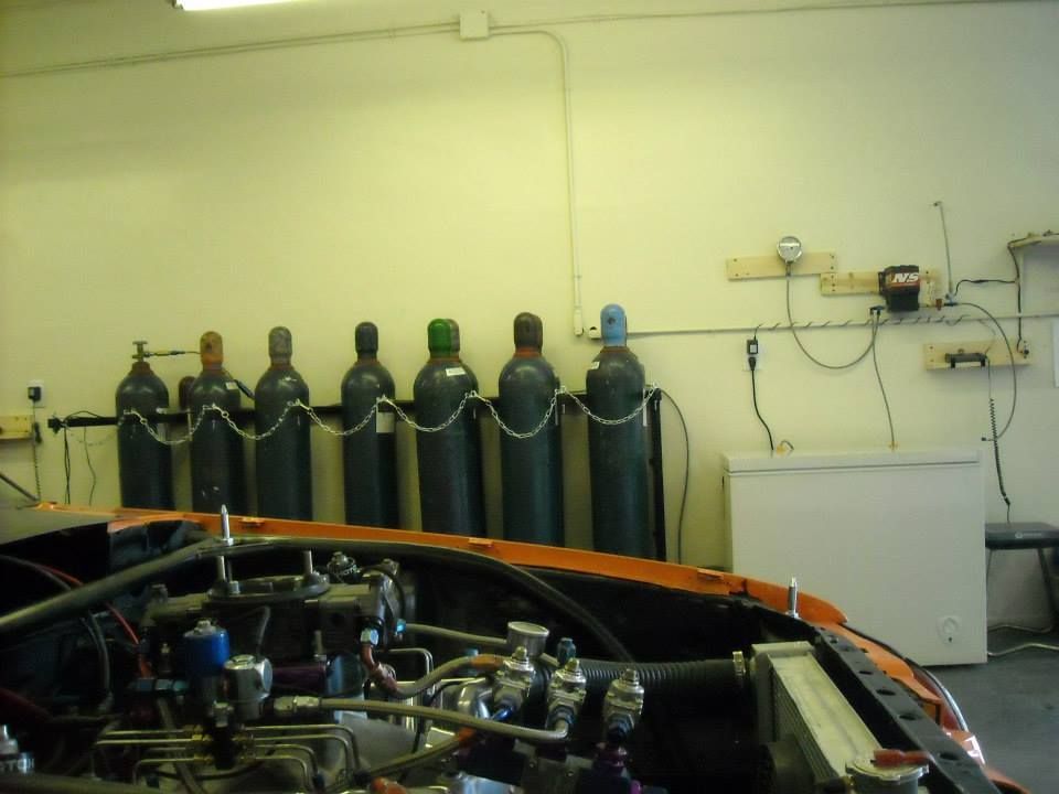 Racing car engine bay in front of a wall with multiple gas cylinders and a small refrigerator.