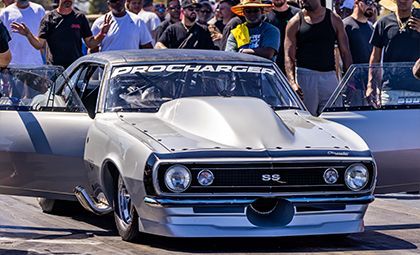 Silver drag racing car with a Procharger logo, at a racetrack, doors open.