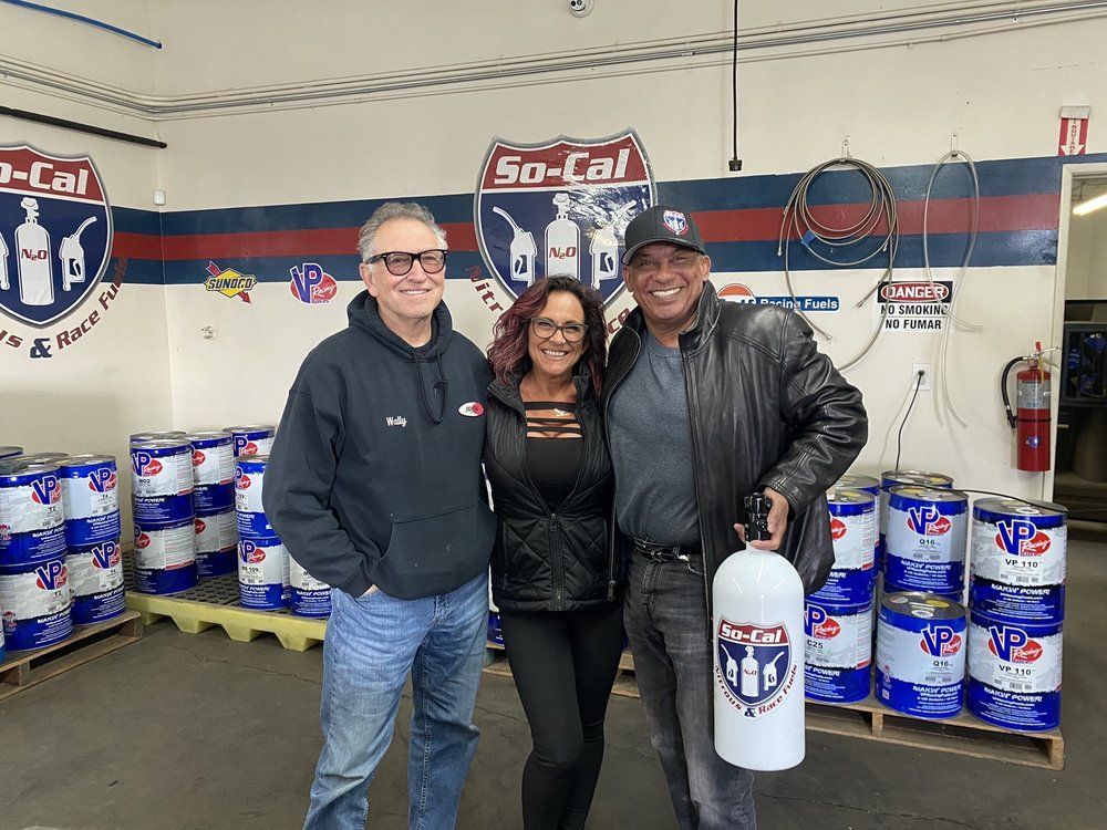 Three people pose in front of stacks of blue and white cans, a So-Cal logo sign in a warehouse.