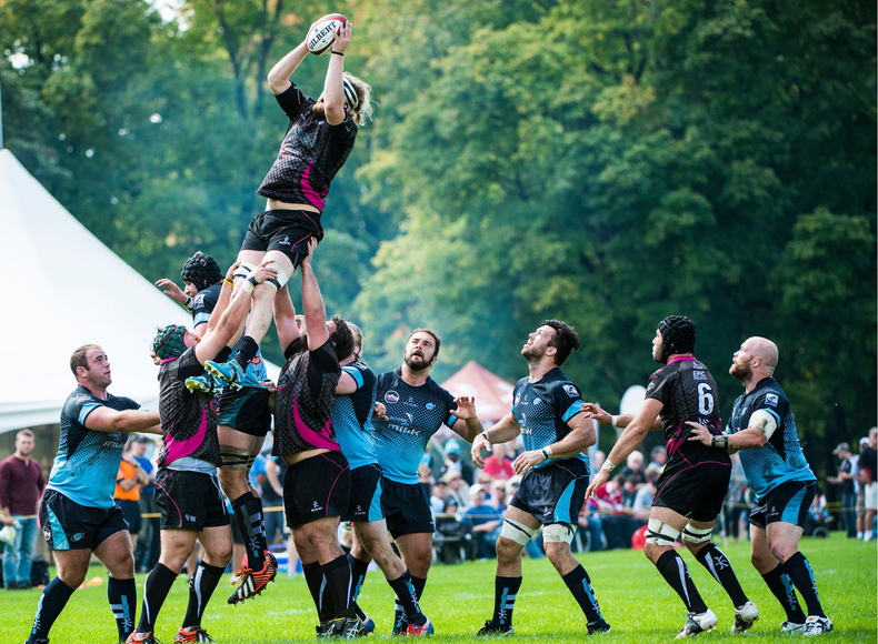 Rugby players in blue and black kits form a lineout, lifting a teammate high in the air to catch the ball on a grassy field.