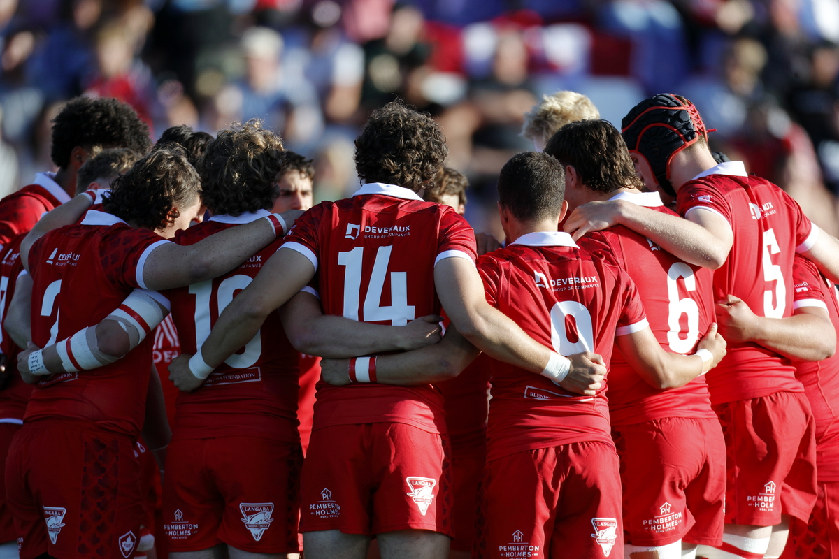 A group of rugby players in red uniforms stand in a huddle on a sunny field, arms around each other's shoulders.