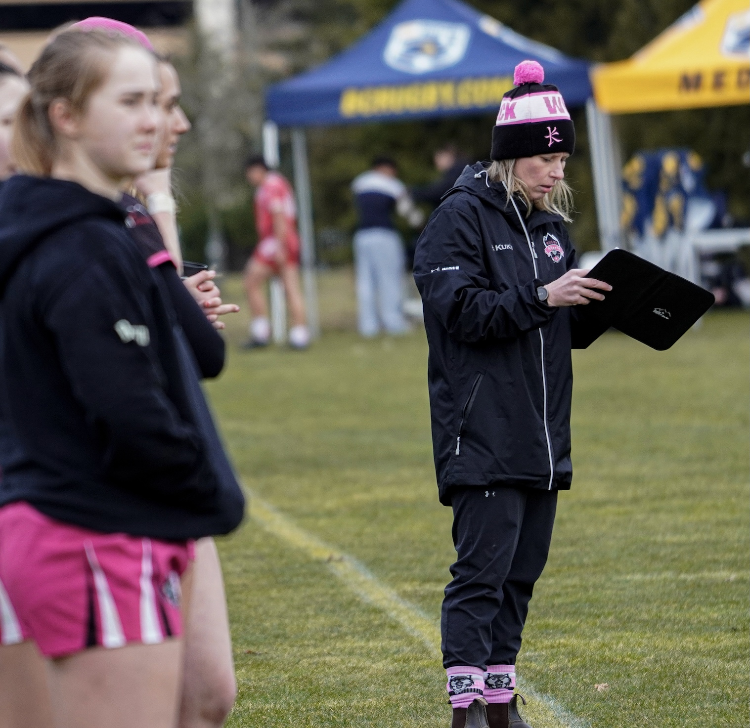 A coach in a black jacket and pink-trimmed beanie reviews notes on a tablet on a sports field near a group of athletes.
