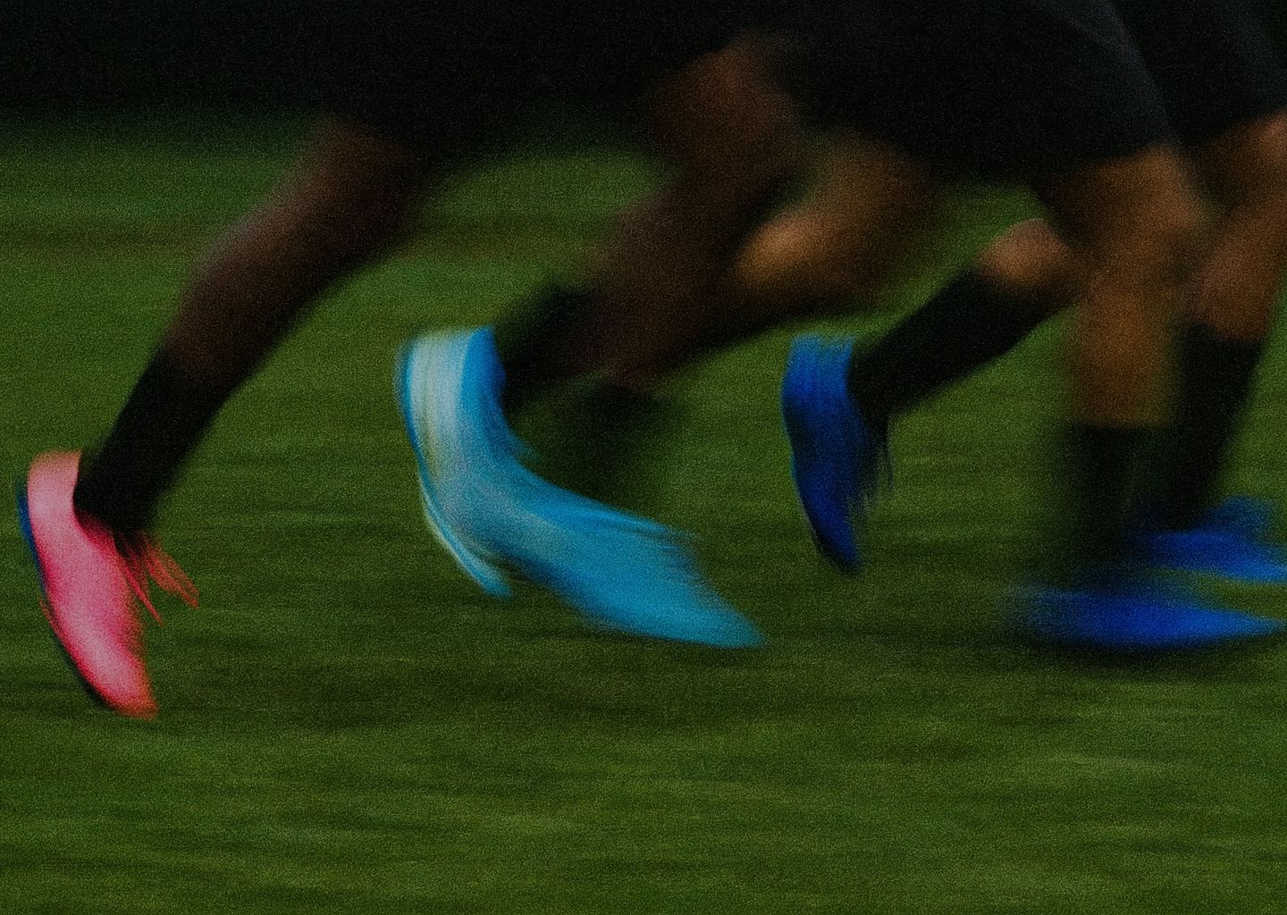 Close-up of soccer players' feet and legs running on a green grass field, wearing vibrant pink and blue cleats.