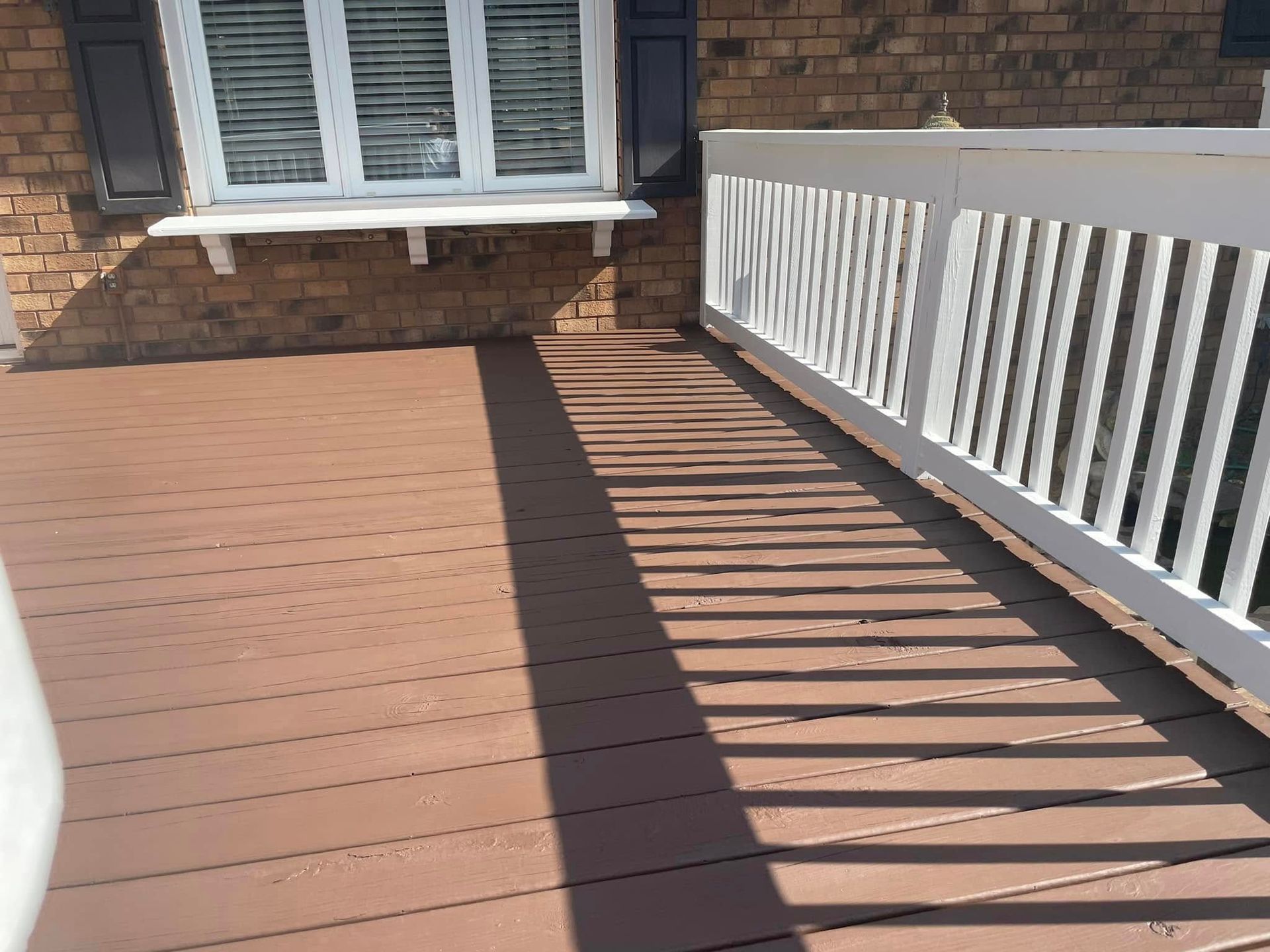 Wooden deck beside a brick house, with white railing casting striped shadows in sunlight.