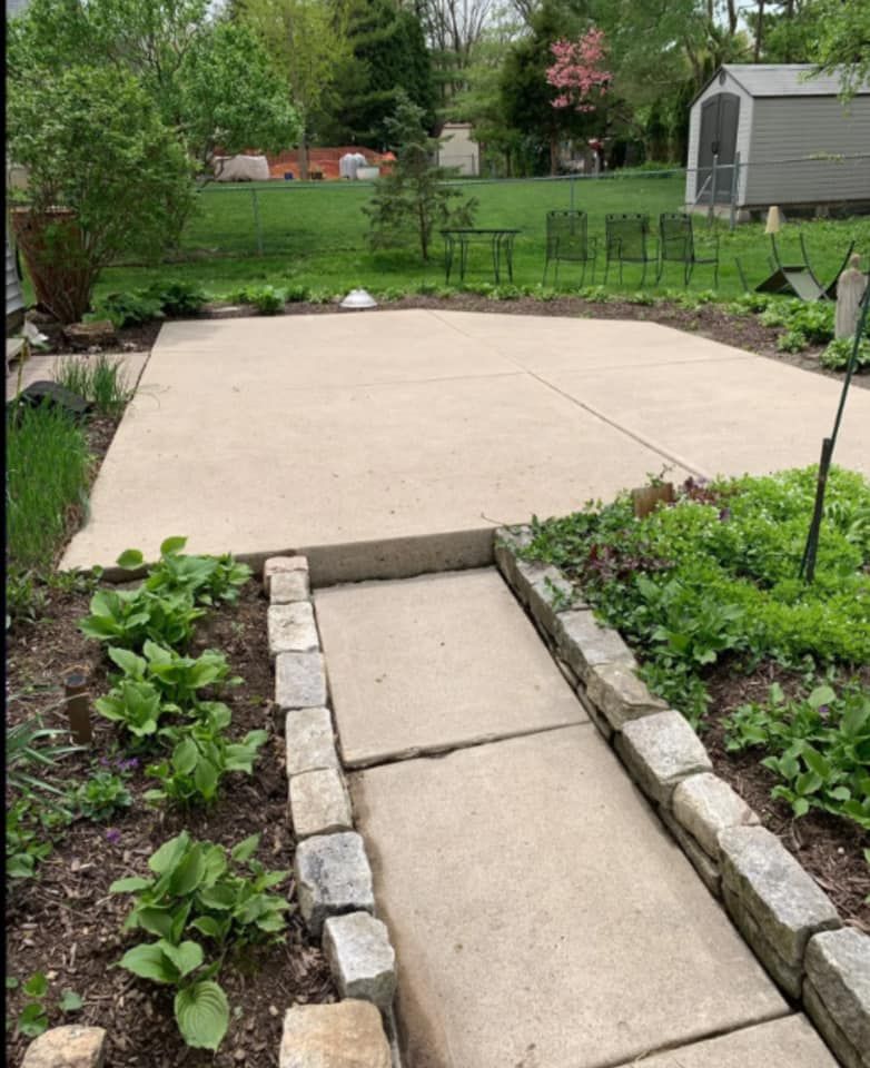 Backyard patio with concrete slab, stone walkway, and garden beds leading to a fenced lawn.
