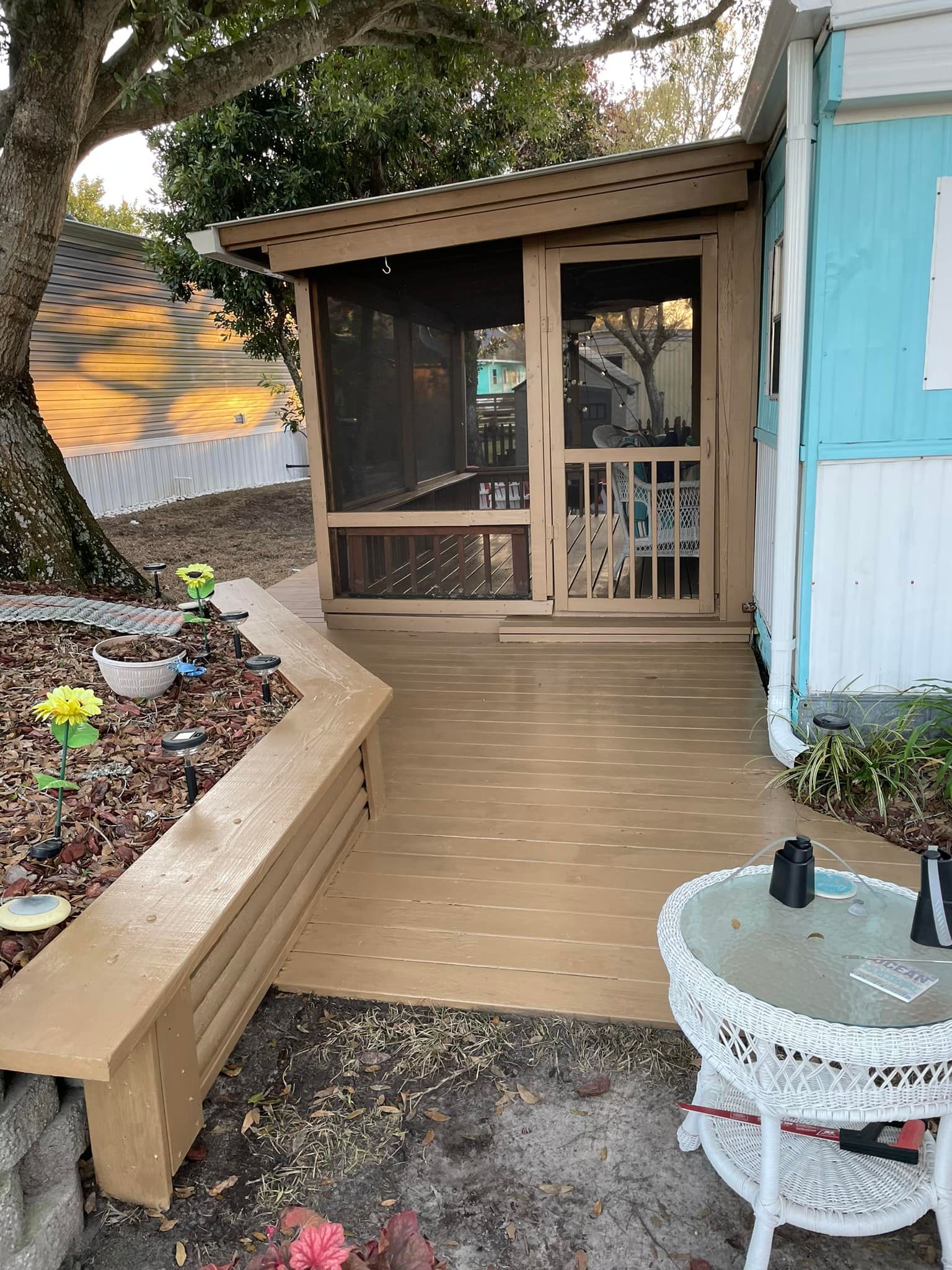 Small porch with screened door, wooden ramp, planter, and a white chair beside a blue exterior wall.