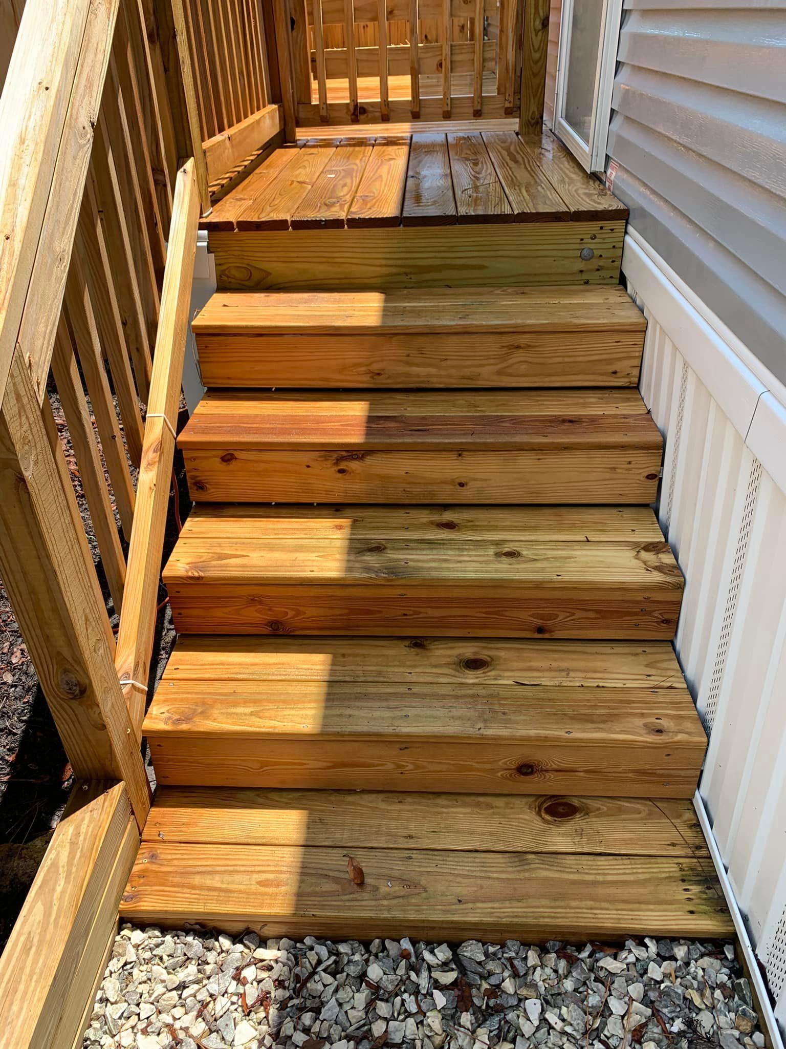 Wooden outdoor stairs leading to a deck, bordered by a white railing and siding, with sunlight and shadows.