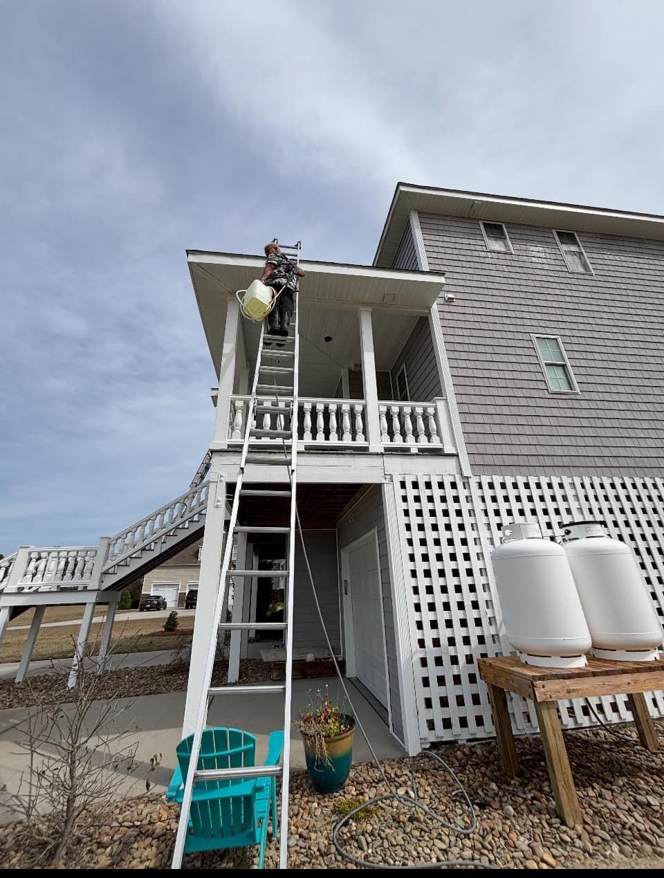 Person on ladder painting upper porch of a gray house, with white railings and water tanks nearby