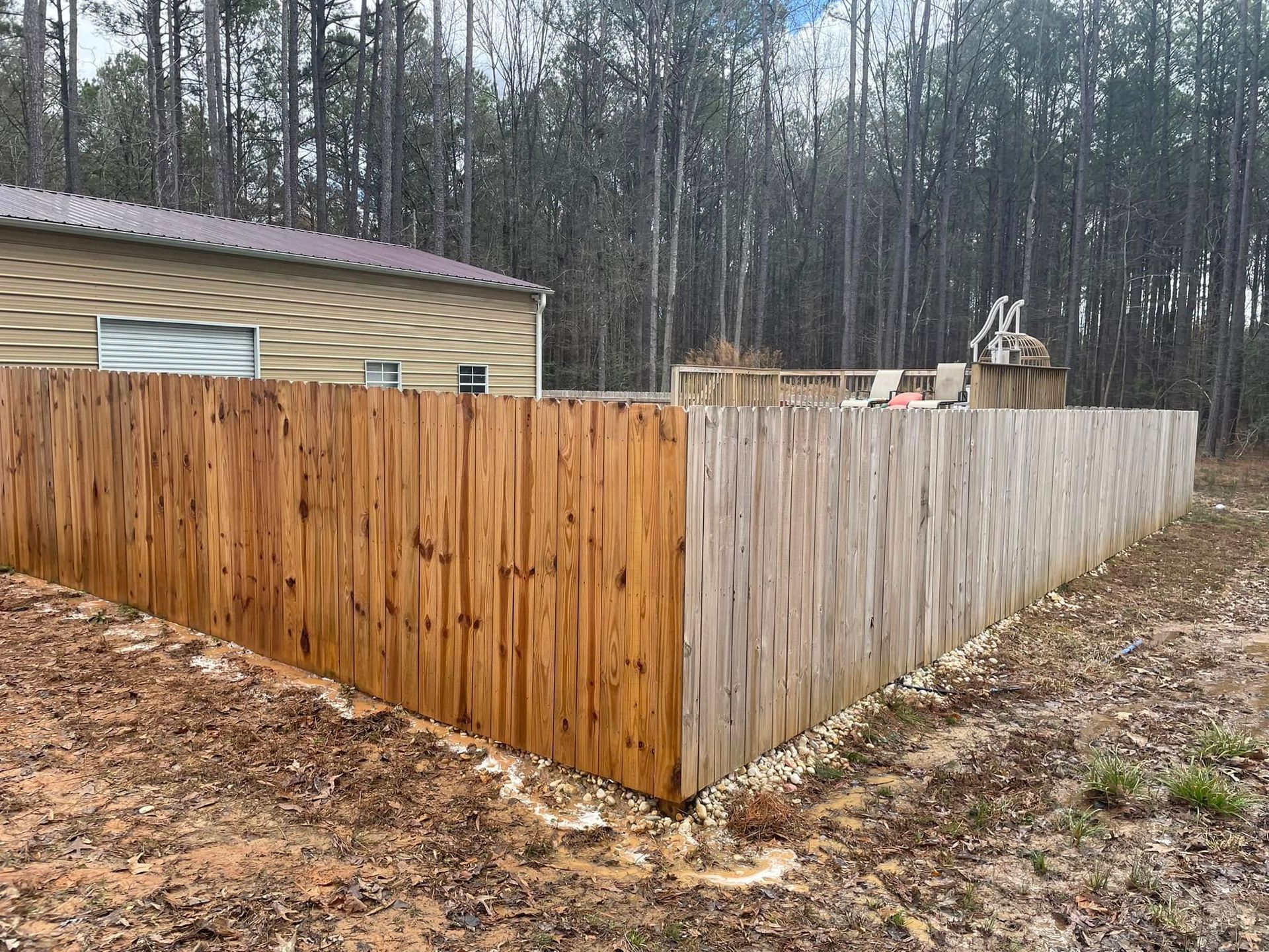 Wooden backyard fence with mixed brown and gray panels beside a beige building and forest trees