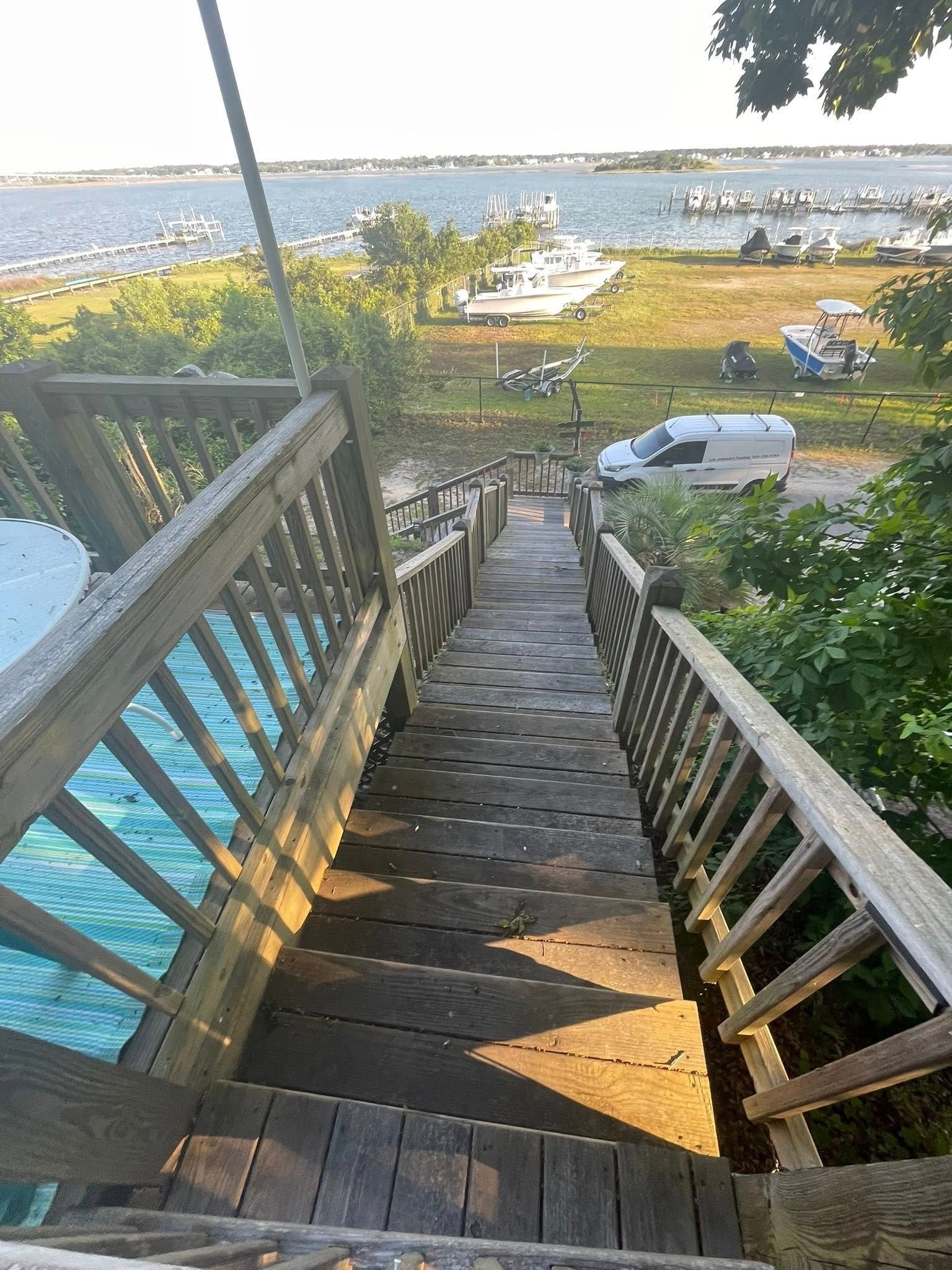 Wooden stairs descend to a lakeside dock with parked cars, boats, and sunlit water beyond.