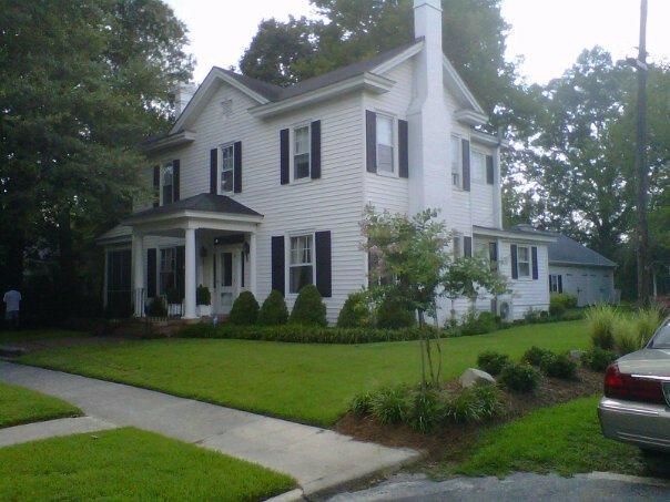 White two-story house with a porch, green lawn, and a driveway in a tree-lined neighborhood.