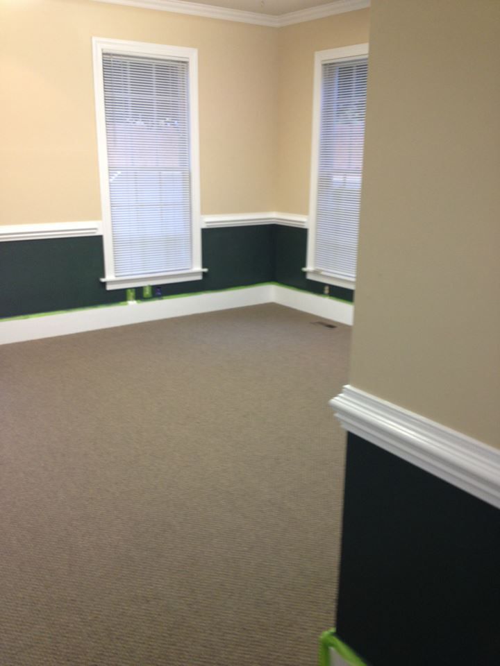 Empty carpeted room with beige walls, dark wainscoting, and two tall windows.