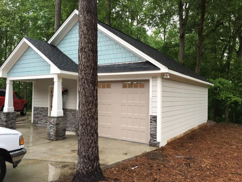 Detached two-car garage with beige siding, blue gable, and stone accents beside a wooded driveway